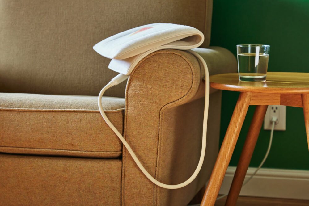 A heating pad folded over the arm of a living room chair with its cord reaching an outlet, and a full glass of water on the side table within arm's reach.