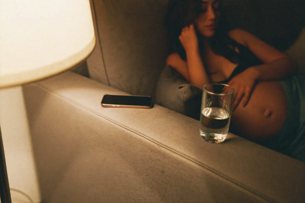 A phone and a glass of water on a sofa armrest in warm evening lamplight, with a pregnant woman resting out of focus behind them.