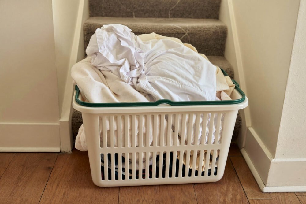 A full white laundry basket heaped with pale laundry, set down on the floor at the foot of a carpeted staircase in a home hallway.