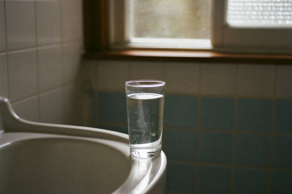 A glass of water on the edge of a bathroom sink in soft morning light