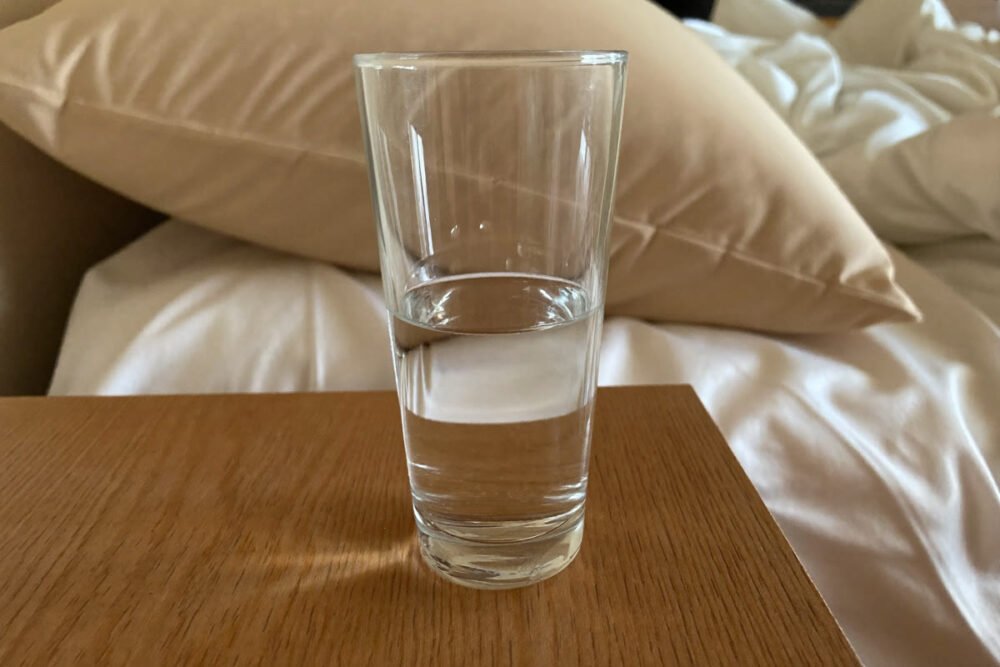 A half-finished glass of water standing on a wooden bedside table in morning daylight, with a rumpled unmade bed out of focus behind it.