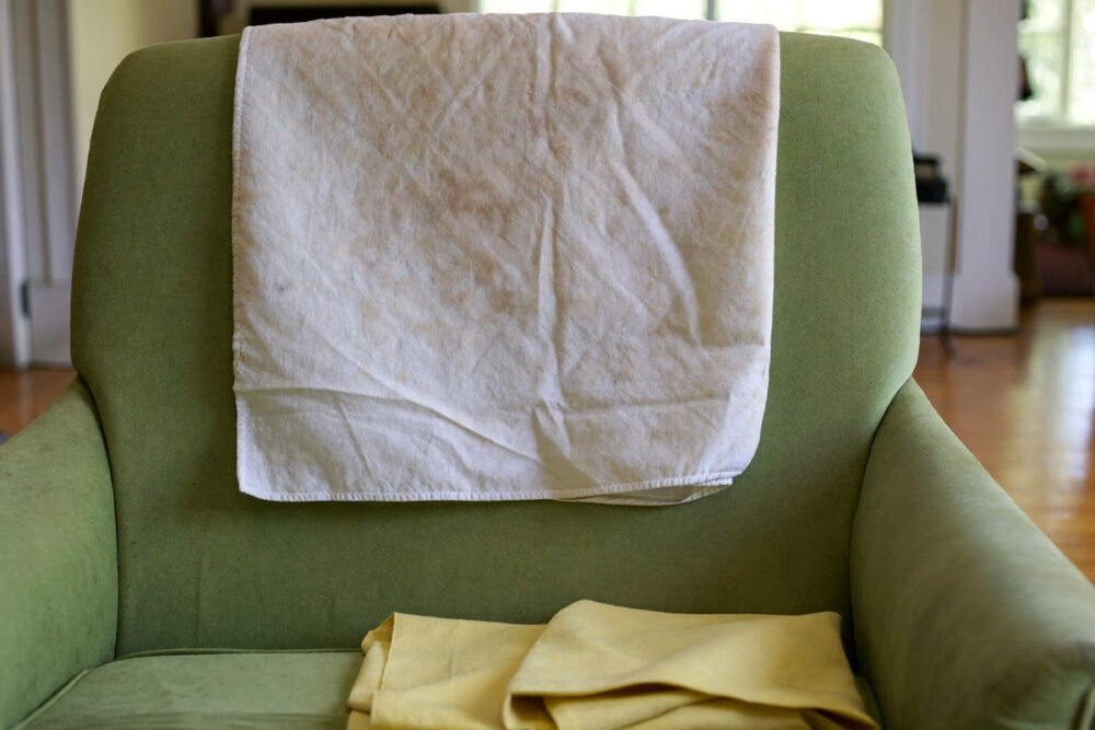 Photograph of a stained white burp cloth draped over the back of a green armchair, with a folded yellow cloth on the seat.