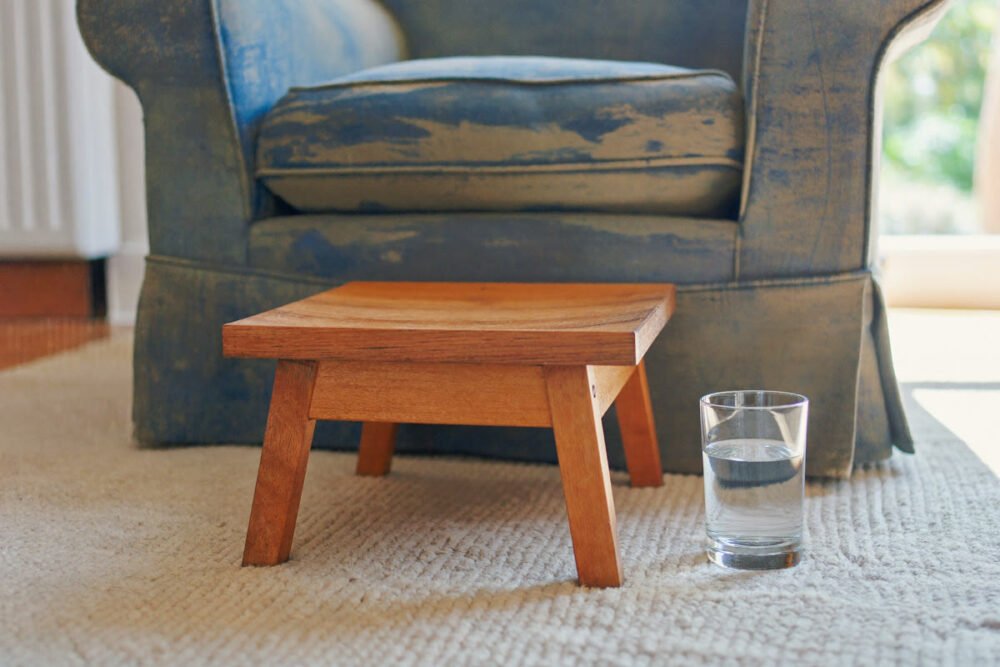 A low wooden footstool on a rug in front of an armchair, with a plain glass of water on the floor beside it, in ordinary summer daylight.